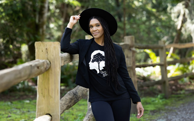 Woman wearing a black shirt and a black hat leaning against a fence with her hand on her hat, posing.